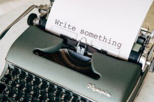Foto: máquina de escrever com folha que tem a frase "write something". Fonte: https://pixabay.com/pt/photos/escrever-autor-um-livro-escrit%C3%B3rio-5243229/